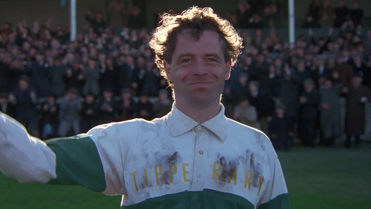 Michael Collins (1996) movie still: medium — An outdoor sports stadium filled with a blurred crowd of spectators sitting in stands. A man in a…