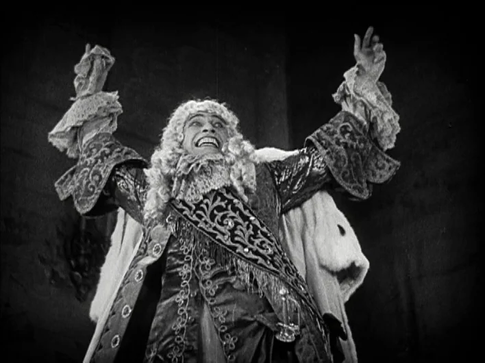 The Man Who Laughs (1928) movie still: medium — An ornate interior features dark, indistinct backgrounds behind a man in historical attire. The man…