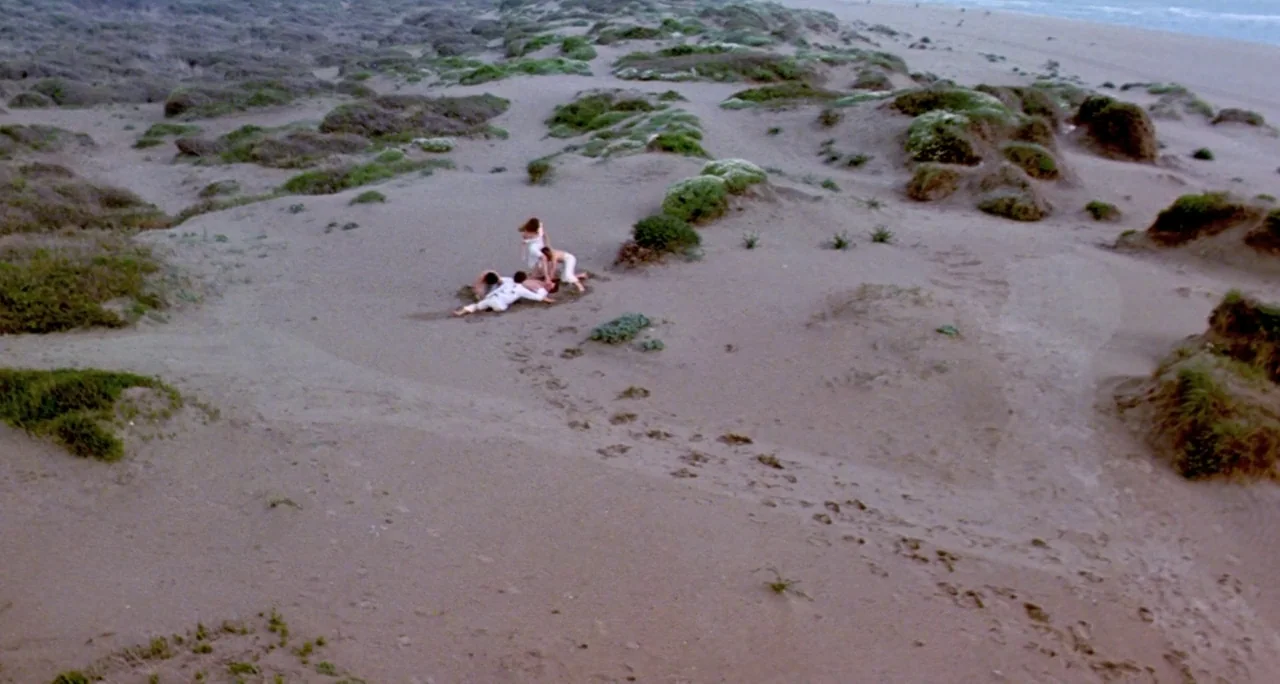 Tenebre (1982) movie still: wide — An expanse of rolling sand dunes covered in low-lying patches of green vegetation. Four individuals in white…
