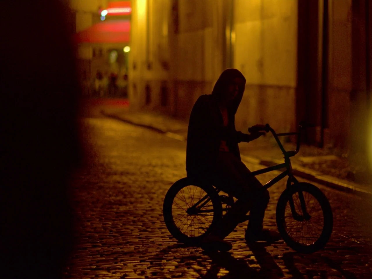 Violet (2014) movie still: medium — A narrow cobblestone street at night is illuminated by a singular orange streetlamp. A hooded figure sits on a…