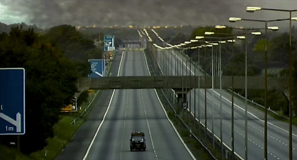 28 Days Later (2002) movie still: wide — A wide multi-lane highway stretches into the distance under a vast, cloud-filled grey sky. A single small…