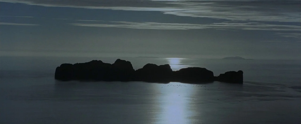 The Beach (2000) movie still: establishing — A dark, jagged island silhouette sits in the center of a calm, moonlit sea. Layers of thin clouds…