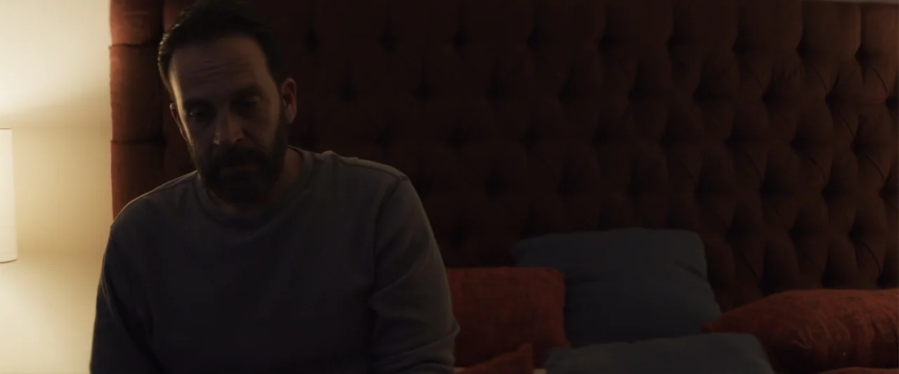 Human Animals (2020) movie still: medium — A dimly lit room with a dark button-tufted headboard behind a person. The man in a grey shirt sits on the…
