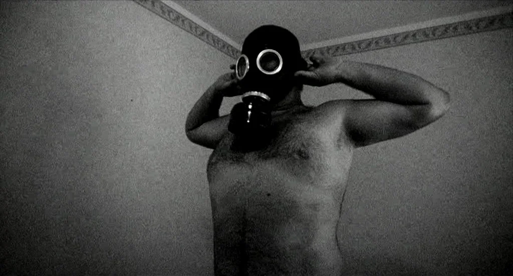 Container (2006) movie still: medium — A corner of a room with plain walls meet under simple decorative molding. A shirtless man stands against the…