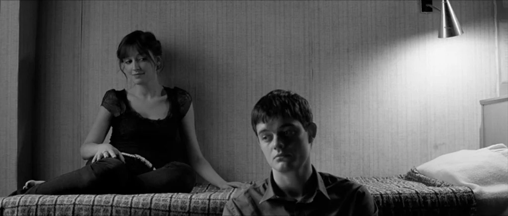 Control movie still: medium — A minimalist bedroom interior features textured wallpaper and a twin bed under a sconce. A man sits facing forward…