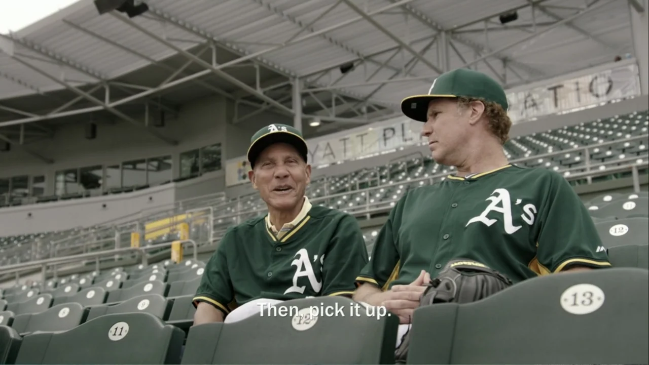 Ferrell Takes the Field (2015) movie still: medium — A set of empty green plastic baseball stadium seats under a large metal roof. Two men in team…