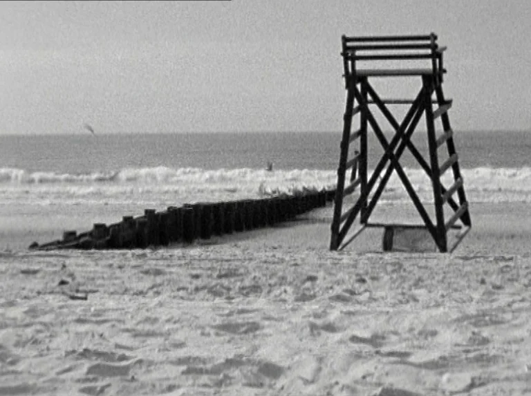 Alice in the Cities (1974) movie still: wide — A wide beach features a wooden lifeguard chair and a long line of pilings. Waves break in the…