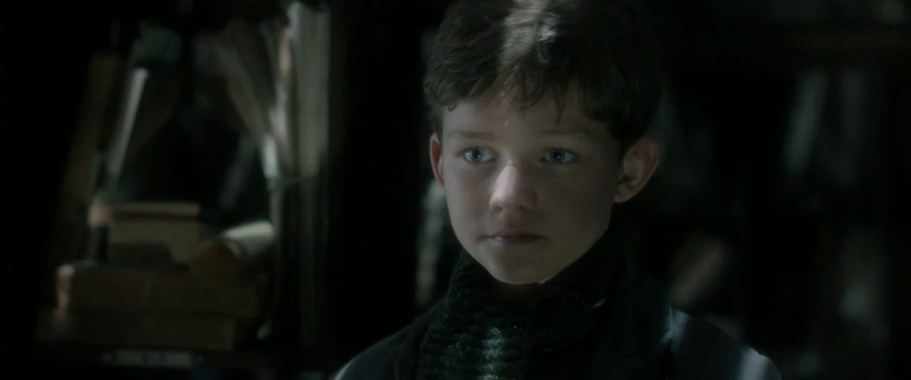 Pan (2015) movie still: closeup — A dimly lit room contains shelves filled with stacks of old paper and books. A young boy with brown hair and a…