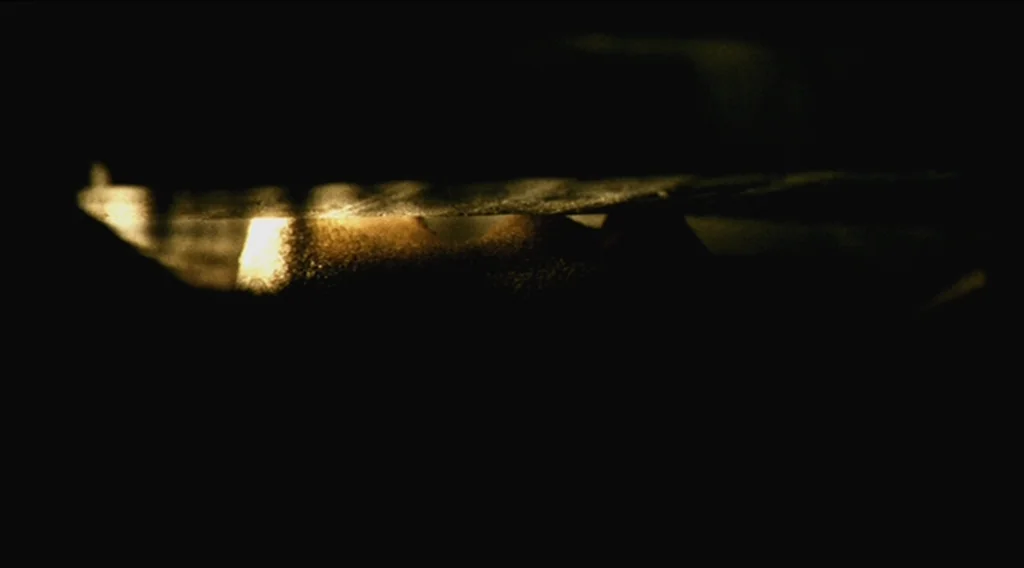 Buried (2010) movie still: closeup — A narrow gap between two dark, opaque objects reveals a patch of warm texture. The frame is largely composed of…
