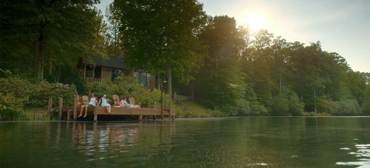 My Best Friend's Exorcism (2022) movie still: wide — A wooden dock extends into a calm lake surrounded by lush green trees. Four young people sit…