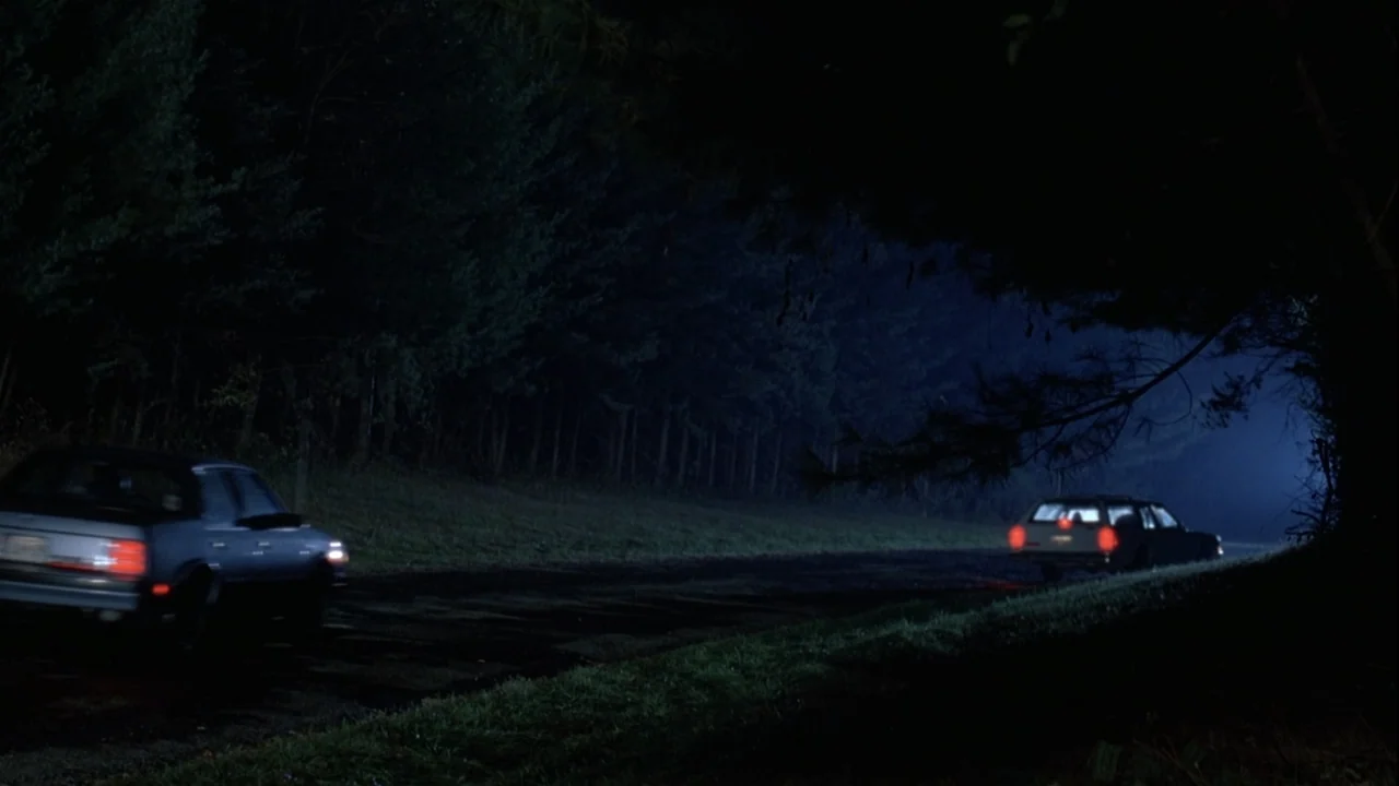 Boys (2021) movie still: wide — A dark, narrow asphalt road winds through a dense forest at night. Two cars with illuminated taillights travel along…