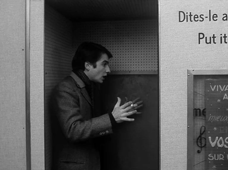Masculin Féminin (1966) movie still: medium — A man stands inside a tight, modular enclosure with a textured pegboard back wall. He gestures with…