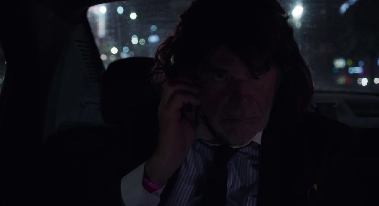 Toni Erdmann (2016) movie still: medium — The interior of a car at night with a blurred cityscape visible through the window. An older man in a suit…