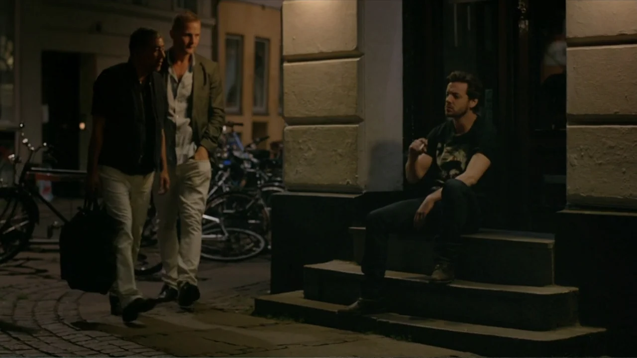 Copenhagen (2002) movie still: fullbody — A stone-paved urban sidewalk sits beside a doorway with heavy plaster wall details at night. Two men walk…