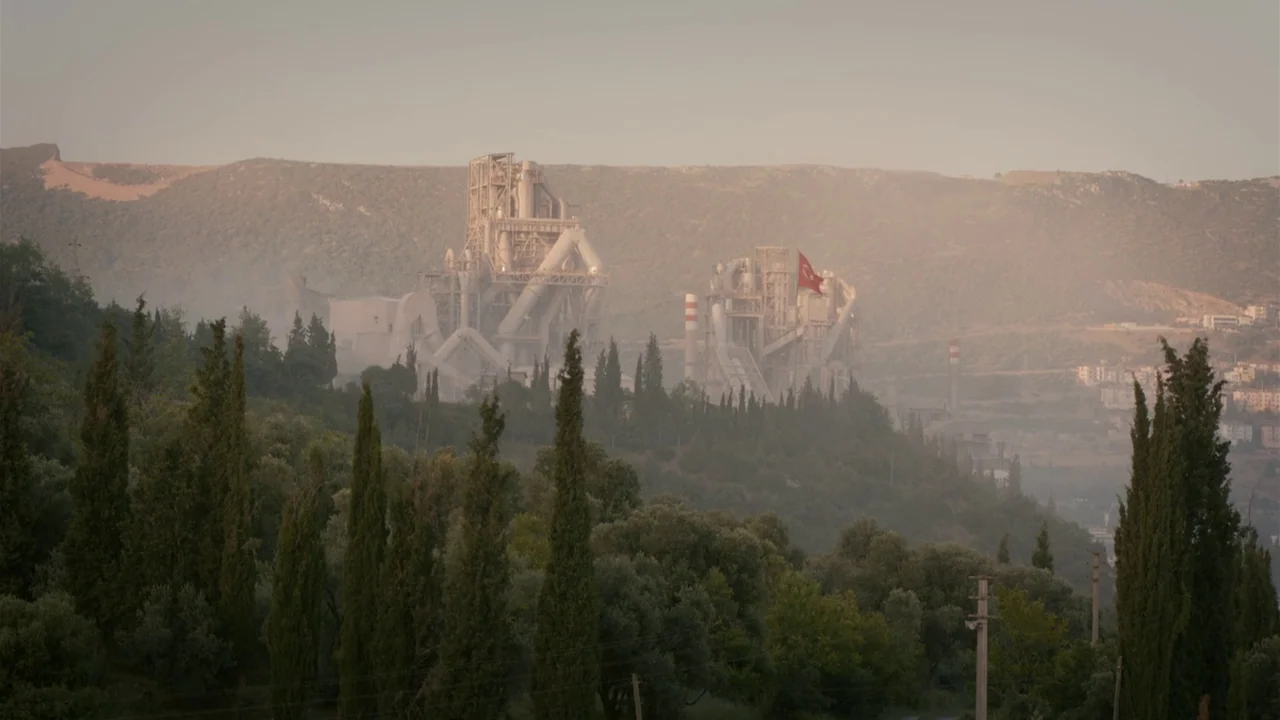 The Field Guide to Evil (2018) movie still: establishing — A large industrial cement factory complex sits nestled in a hilly landscape with…