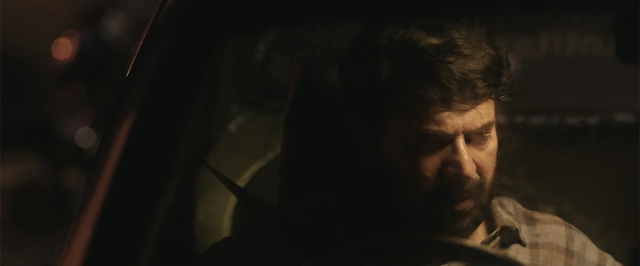 Peranbu (2019) movie still: closeup — A dark vehicle interior with a man visible in the driver seat. The man drives the car while looking down…