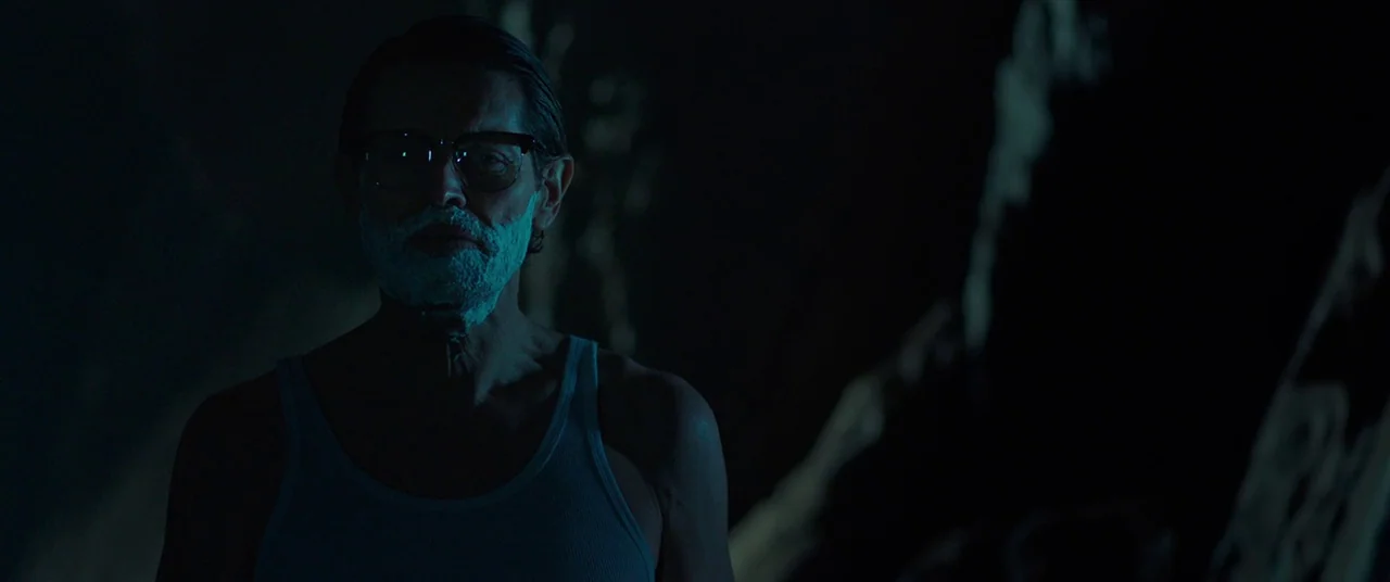 Siberia (2019) movie still: medium — A dimly lit, dark space features a man with white foam on his beard. The man wears a white undershirt and…