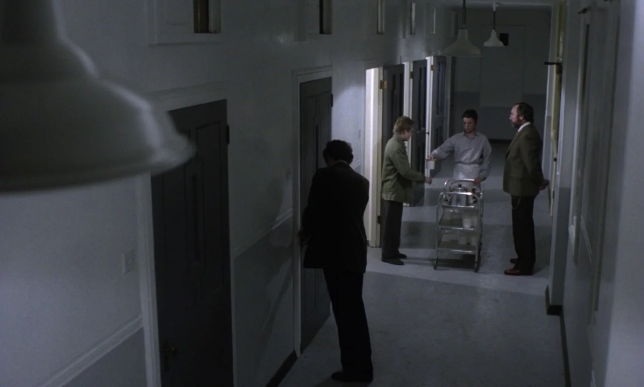 Scum (1979) movie still: wide — A long, dimly lit institutional corridor with multiple closed doors along both walls. A man in a suit approaches a…