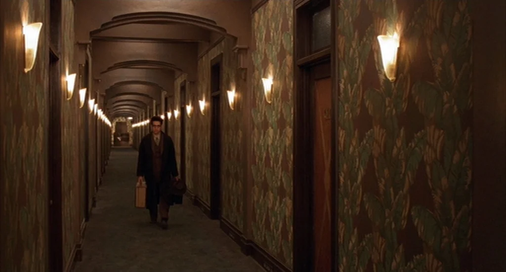 Barton Fink (1991) movie still: fullbody — A long, narrow corridor features vintage wallpaper with green leaf patterns and arched ceilings. A…