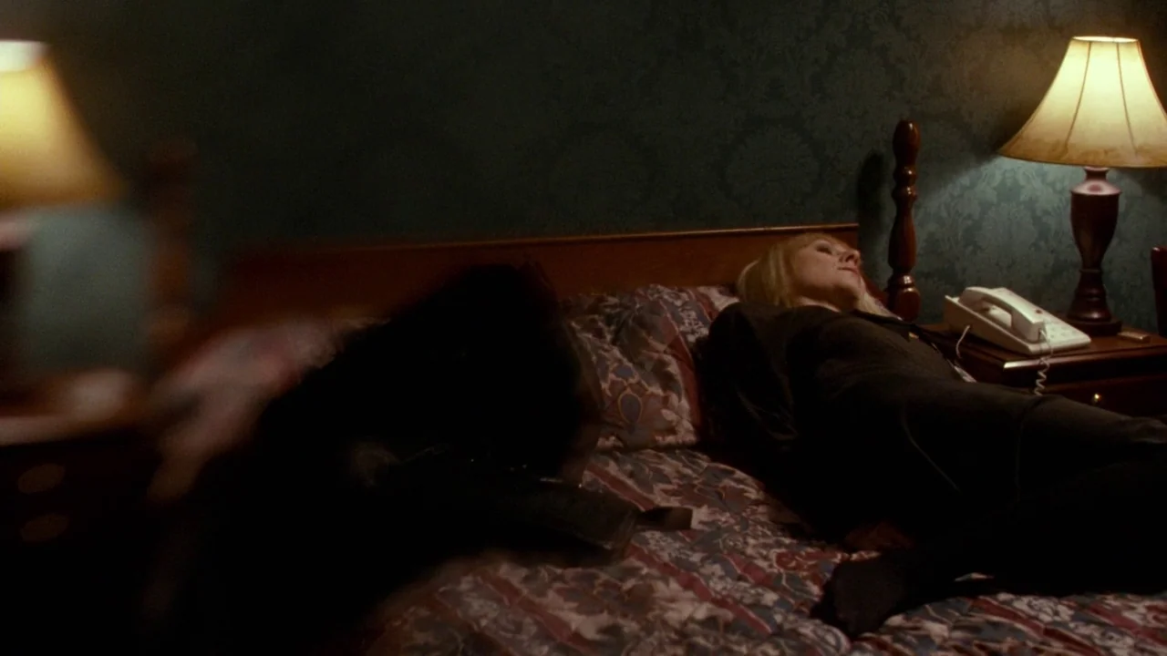 Cold Souls (2009) movie still: medium — A dimly lit hotel room features dark green patterned wallpaper and a wooden headboard. One woman lies on the…