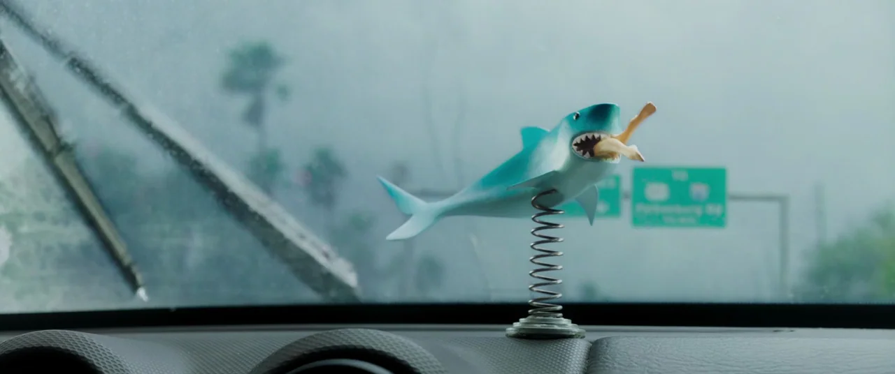 Crawl (2019) movie still: closeup — A plastic shark toy on a coil spring sits atop a vehicle dashboard. Through the rainy windshield, a blurred…