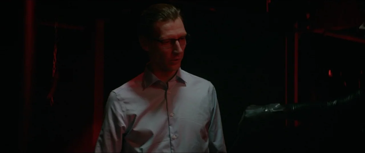 Dogs Don't Wear Pants (2019) movie still: medium — A dark, minimally furnished interior space is bathed in a distinct red light source. A man in a…