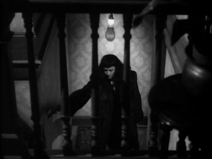 The Seventh Victim (1943) movie still: medium — A woman in a dark coat descends stairs past ornate wooden banisters. A single bare light bulb…