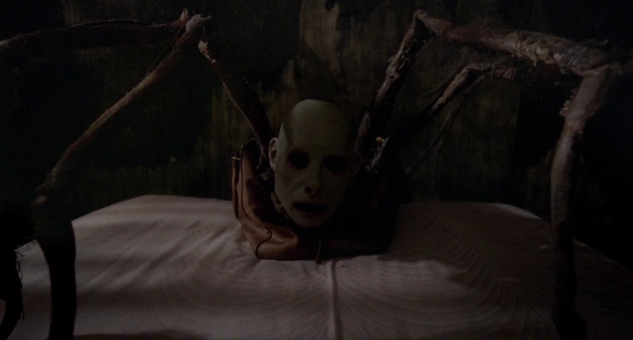 Possum (2018) movie still: medium — A dark, nondescript room features a pale-headed creature with spider-like limbs. The entity sits atop a white,…