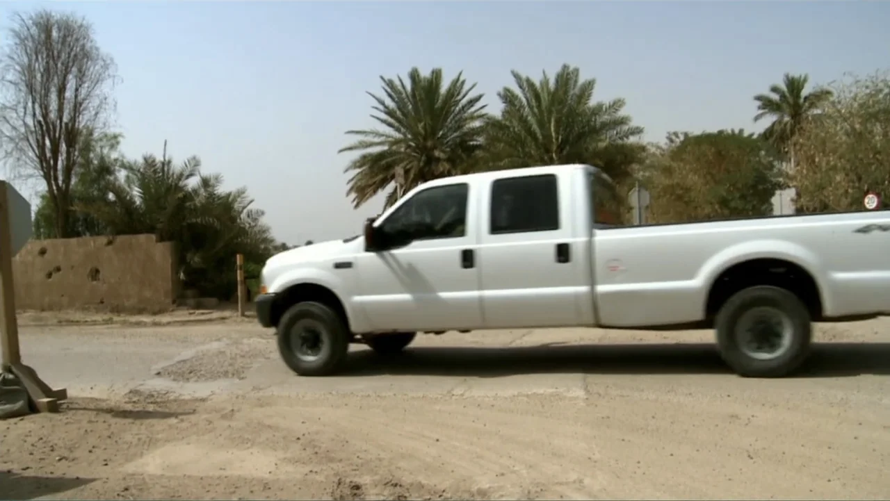Wartorn: 1861-2010 (2010) movie still: medium — A white pickup truck moves across a dusty road flanked by sparse desert vegetation. Bright daylight…