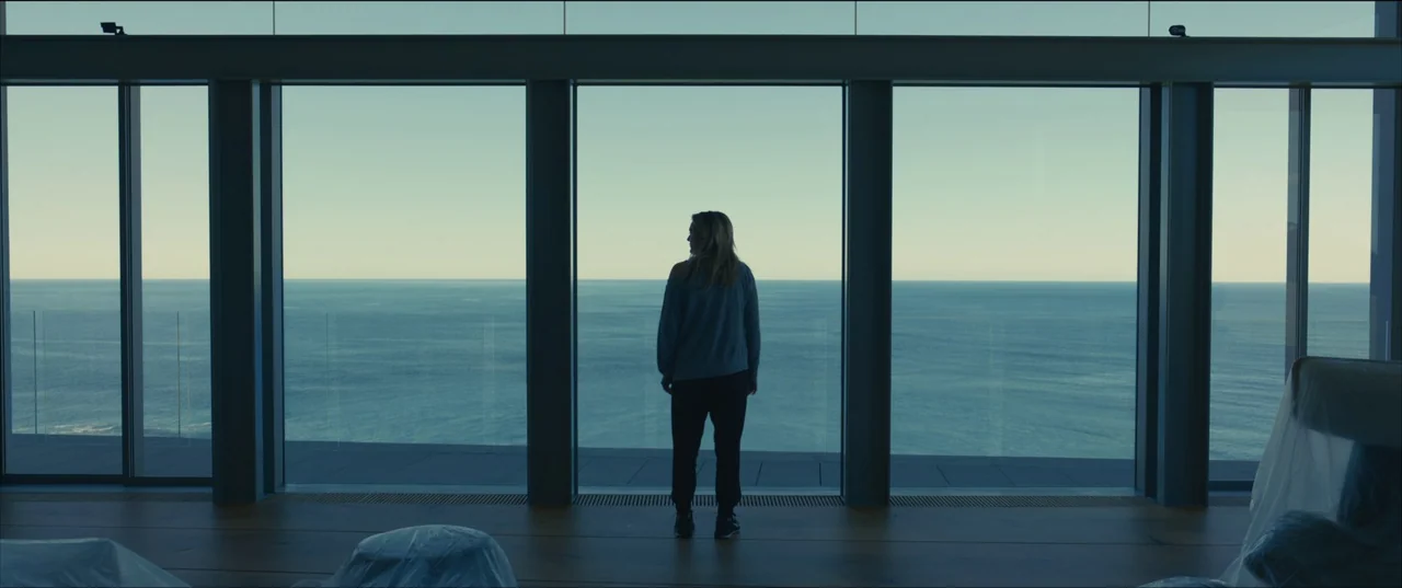 The Invisible Man (2020) (2020) movie still: fullbody — A spacious room features floor-to-ceiling glass windows overlooking a calm sea. A woman…
