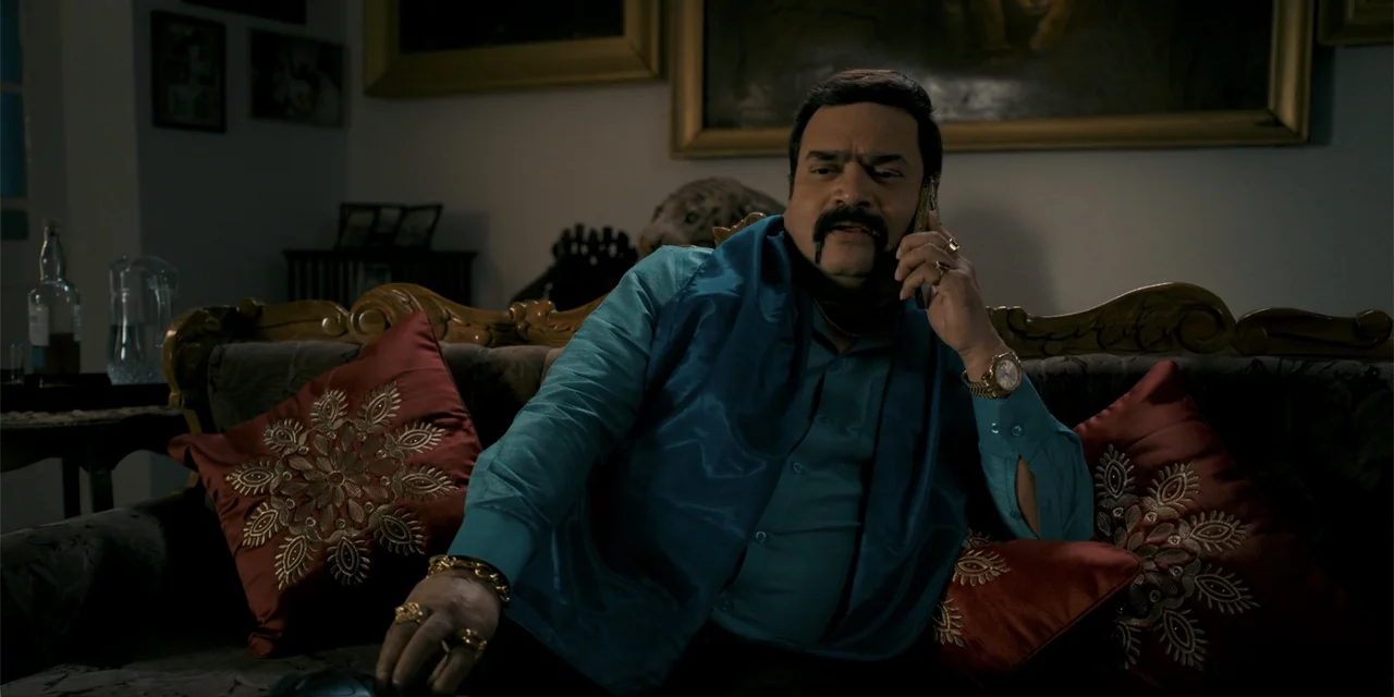 Dry Day (2023) movie still: medium — A dimly lit room features a dark sofa with red decorative cushions and framed artwork. A middle-aged man sits…