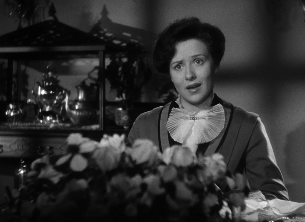 Citizen Kane (1941) movie still: medium — A dimly lit room features a decorative glass case displaying silver tea service items. A woman wearing a…