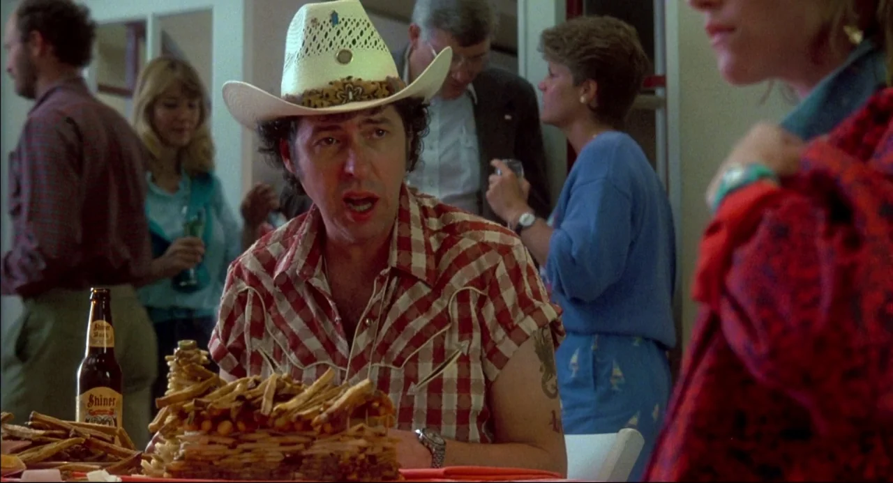 The Texas Chainsaw Massacre 2 (1986) movie still: medium — A man in a cowboy hat sits at a table during a social gathering. He is positioned in…