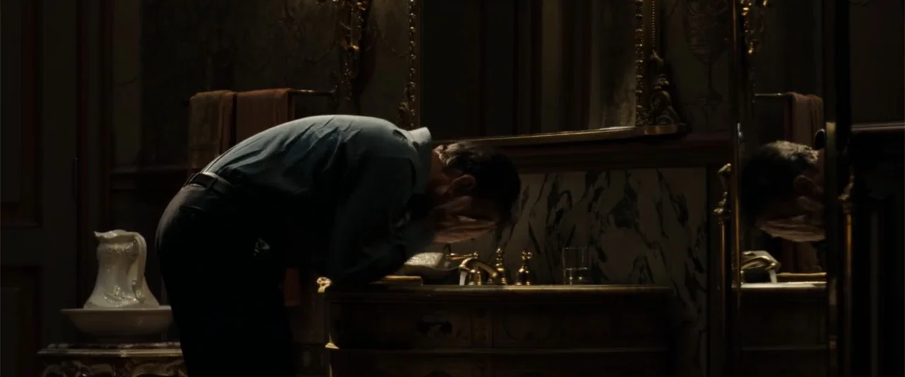 Angels & Demons (2009) movie still: medium — A dark bathroom features a marble-topped vanity with an ornate mirror and fixtures. A man stands at the…