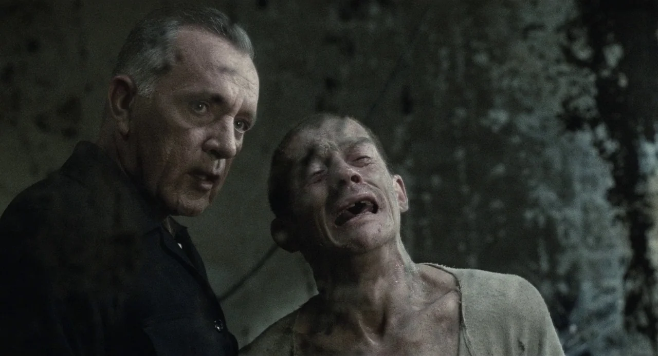 1984 (1984) movie still: medium — A dimly lit, concrete-walled room shows signs of age and industrial decay. An older man observes a distressed,…