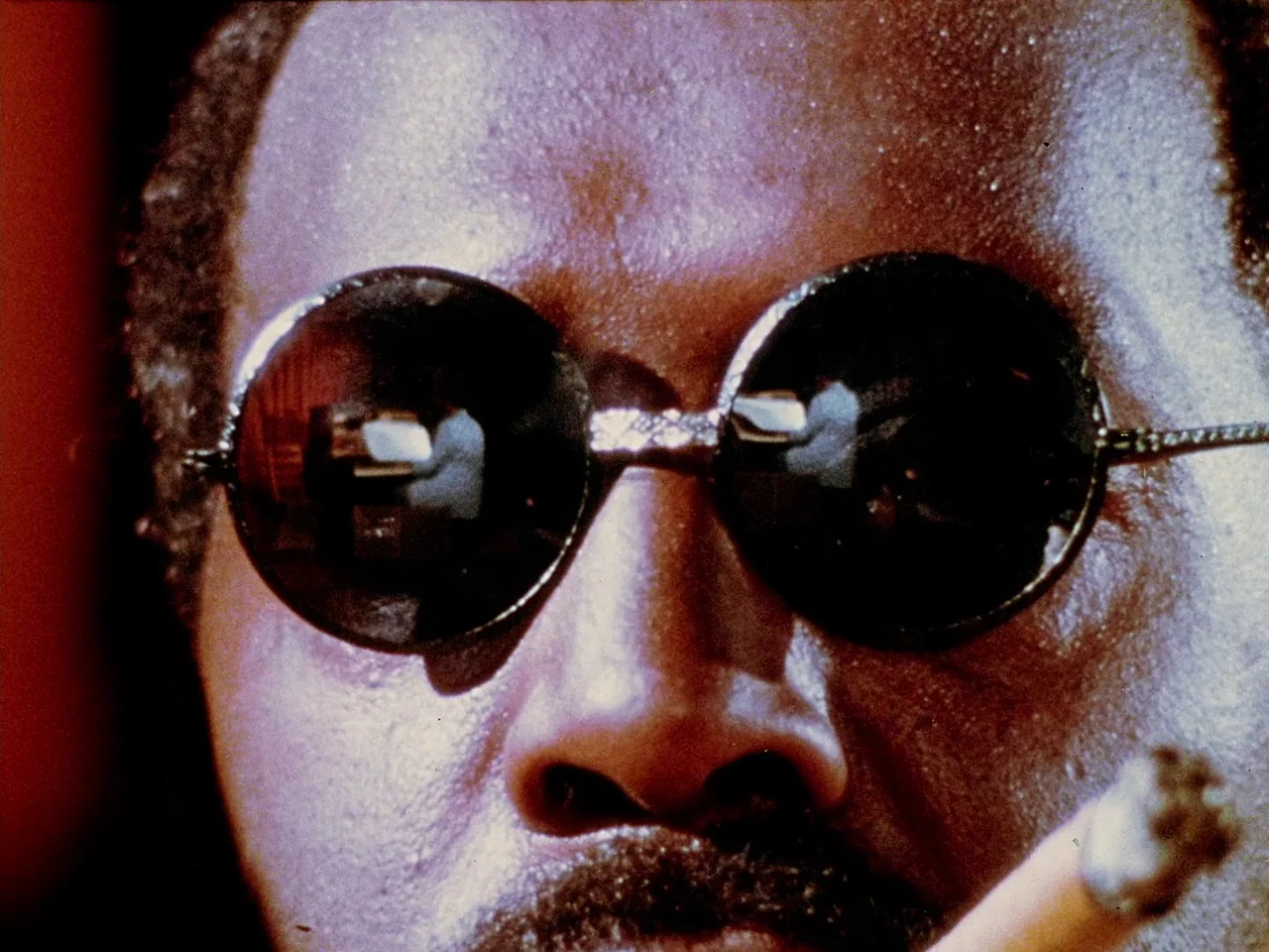 Space is the Place (1974) movie still: closeup — A close-up of a man wearing large round dark sunglasses with visible reflections. The man holds a…