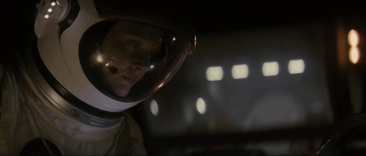 The Last Days On Mars (2013) movie still: closeup — An astronaut wearing a white helmet is seated inside a dark cockpit cabin. The interior is dim…