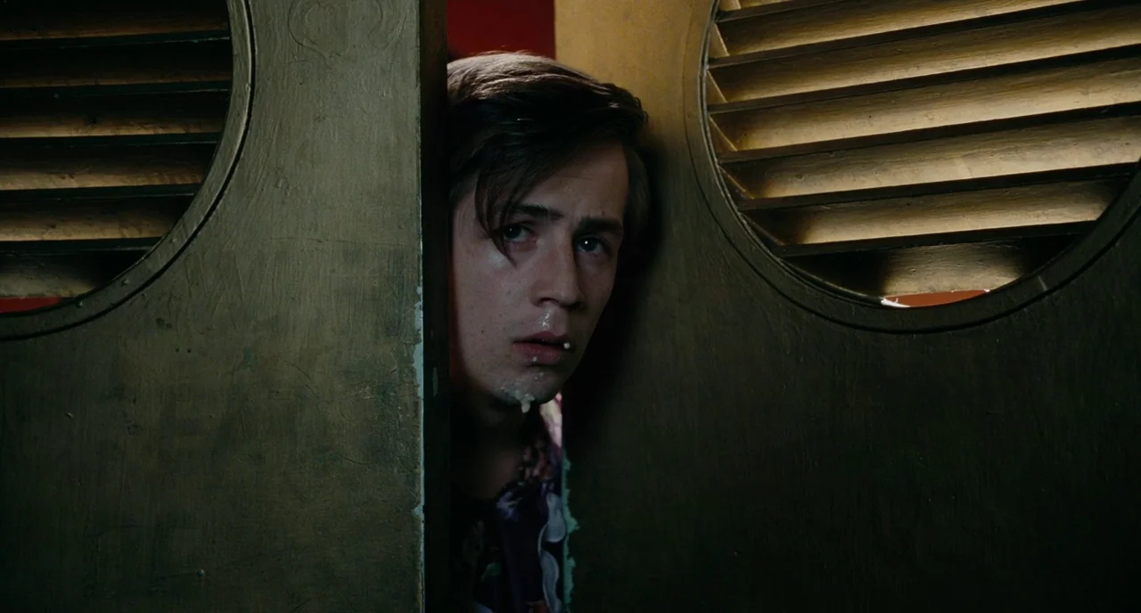 Gentlemen Broncos (2009) movie still: closeup — A man peeks through a narrow gap between two dark wooden saloon doors. The subject shows a surprised…