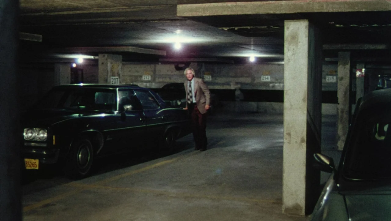 Shivers (1975) movie still: wide — A dimly lit concrete parking garage features numbered stall markers painted on the rear walls. An older man in a…