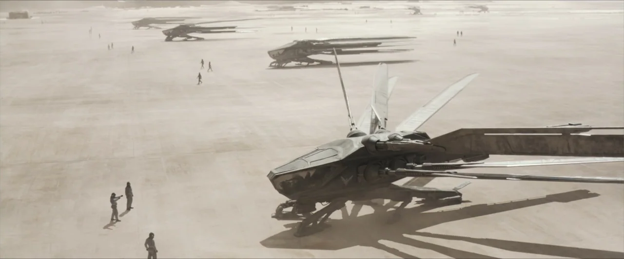 Dune (2021) movie still: establishing — A vast, flat desert landing strip features multiple parked advanced aircraft under bright daylight. Several…