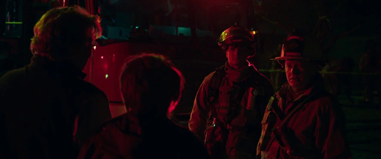Dark Waters (2019) movie still: medium — A nighttime scene features firefighters standing near a vehicle marked with emergency red lights. They…