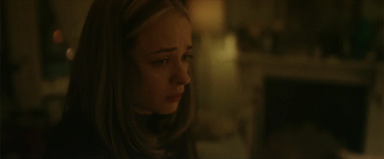 Better Man (2024) movie still: closeup — A dimly lit room features a blurred fireplace hearth in the background. A young woman with light brown hair…