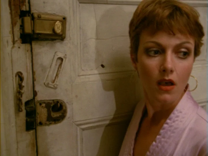 Tenement (1985) movie still: closeup — A close-up of a woman stands next to a white wooden door with metallic locks. The door has visible cracks and…