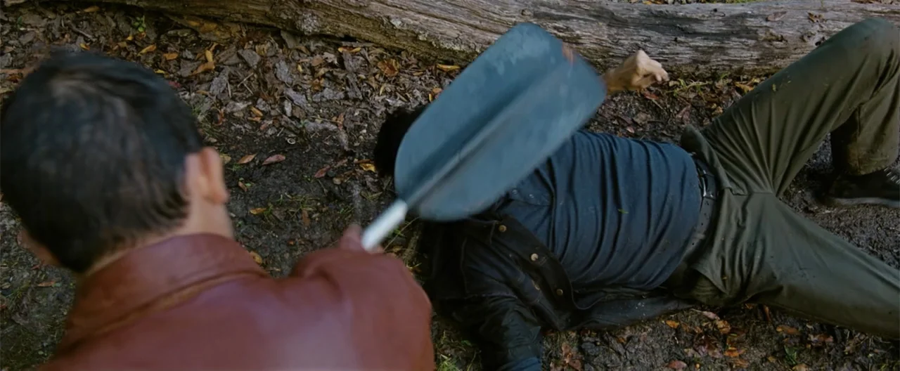 Camp Hideout (2023) movie still: medium — A man in a leather jacket holds an oar over a prone person outdoors. The ground is covered in dirt and…