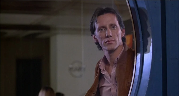 Videodrome (1983) movie still: medium — A man stands in a doorway looking into a darkened interior space with wall signage. The man faces the camera…