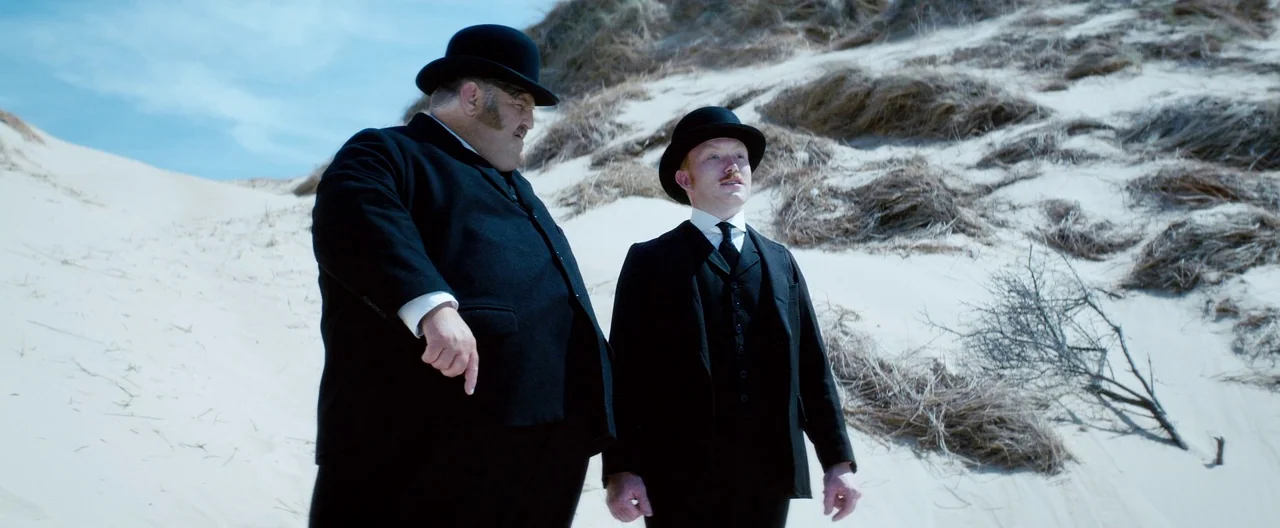 Slack Bay (2016) movie still: fullbody — Expansive white sand dunes with tufts of dry grass populate a sunny coastal landscape. Two men wearing…
