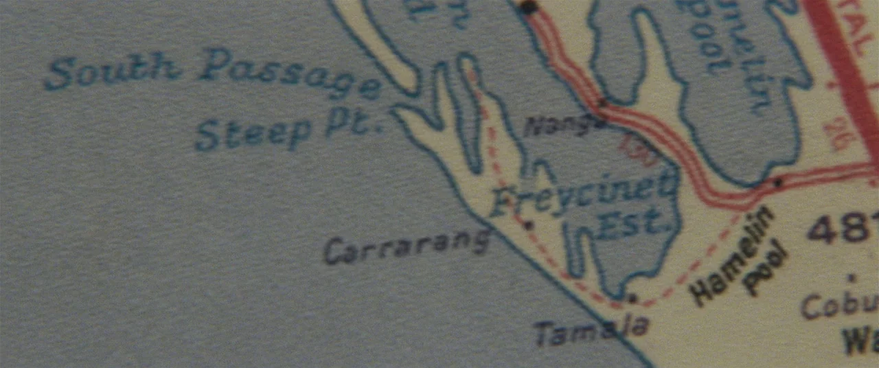 Tracks (2013) movie still: closeup — A close-up view of a printed geographical map with blue coastal regions. Text labels and red dashed routes are…