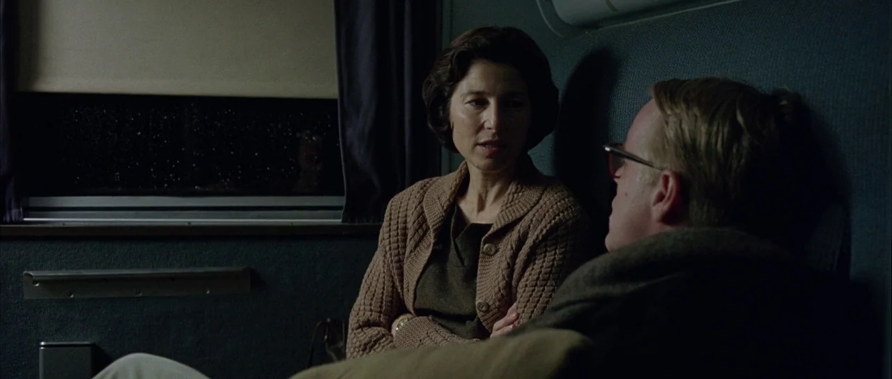 Capote (2005) movie still: medium — A dimly lit train compartment features blue-grey upholstery and a dark window at night. A woman in a brown…