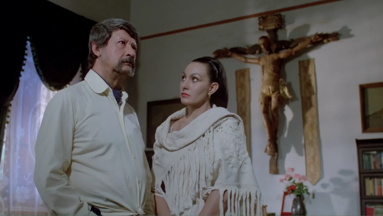 Toña Machetes (1985) movie still: medium — A domestic interior features a white wall adorned with a large wooden crucifix. An older man and a…
