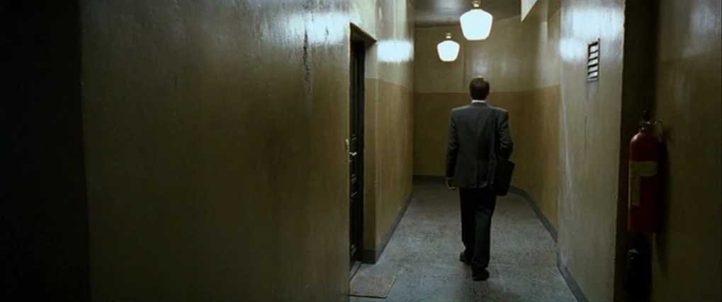 Next Door (2005) movie still: fullbody — A narrow apartment building corridor features ochre walls and a stone tiled floor. A man in a dark suit…